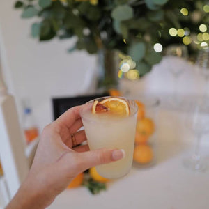 A hand holding a frosty margarita garnished with a slice of orange, with a blurred background featuring a vase of greenery and a festive setting with string lights.