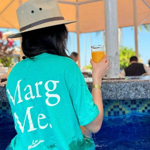 A person wearing a hat and a teal 'Marg Me.' T-shirt, relaxing at a poolside bar with a drink in hand, enjoying the sunny outdoor setting under a canopy