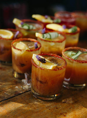 A close-up of several glasses filled with a spicy margarita, garnished with lemon slices, cucumber ribbons, and small purple flowers, served with tajin-rimmed edges on a rustic wooden surface