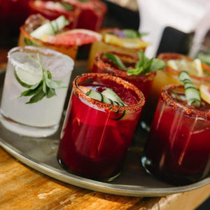 A tray of assorted colorful margaritas, with fresh herbs, cucumber slices, and citrus, served in glasses with tajin on the rim, creating a vibrant and refreshing display.