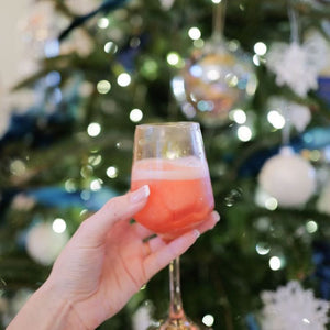 A hand holding a wine mix drink, with a blurred background featuring a vase of greenery and a festive setting with string lights.