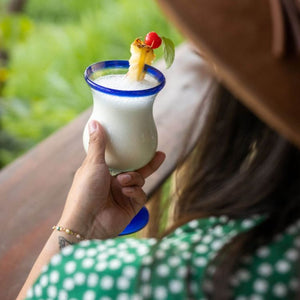 A person holding a creamy piña colada in a blue-rimmed glass, garnished with a pineapple slice, cherry, and mint leaf, against a lush green outdoor background