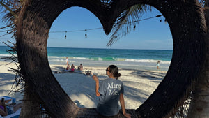 A person wearing a 'Marg Me.' T-shirt sits within a large, heart-shaped wicker frame, enjoying a drink while looking out at the beach and ocean, with a few people relaxing on the sand in the background under a clear blue sky.